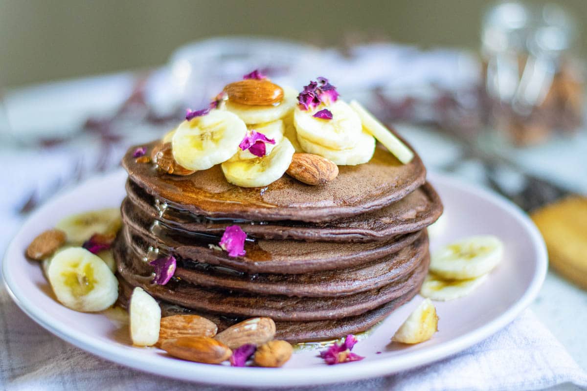 chocolate banana pancakes on a plate topped with almonds, bananas, and pink petals