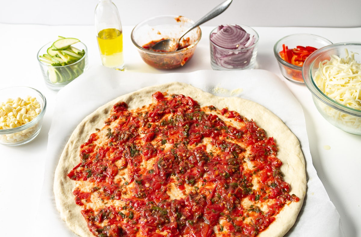 pizza crust covered with Vegan Pizza Sauce and surrounded by bowls of toppings