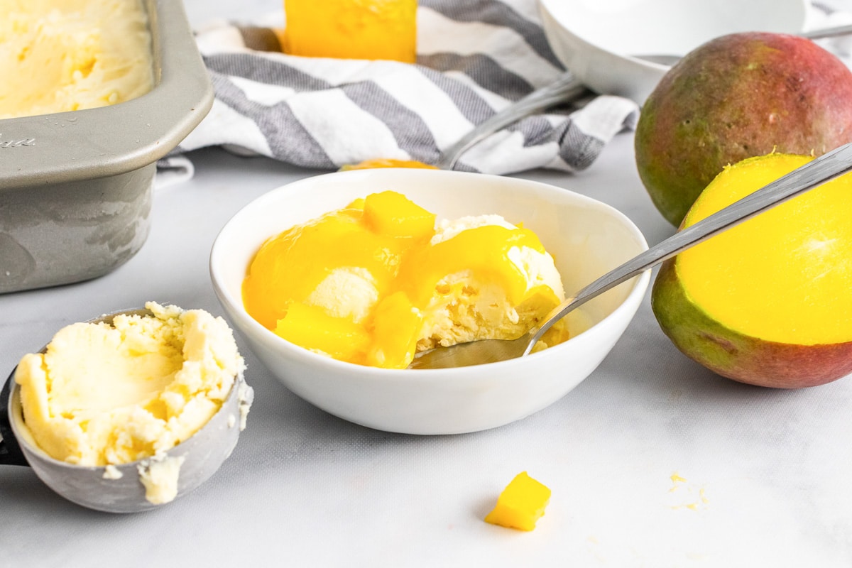 bowl of homemade mango ice cream covered in puree and surrounded by mangoes on table